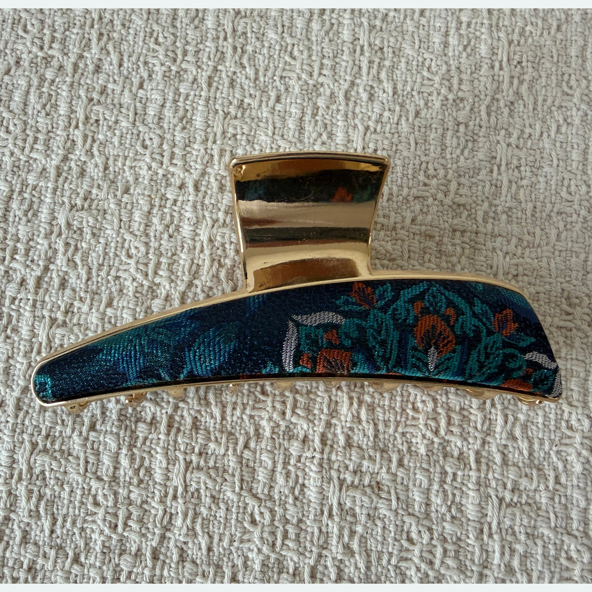 Teal Floral Curved Bar Hair Claw Clip by JOFAE Kyoto. Tapered curved bar silhouette; Dark teal floral textile detail; Turquoise and orange accents; Gold-tone claw frame.
